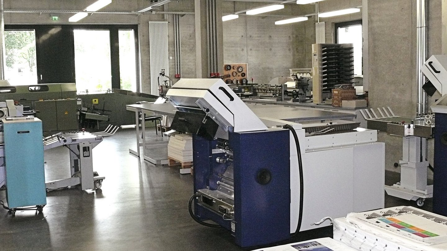Maschinensaal des Bedruckstoffverarbeitungslabors mit Falzmaschine im Vordergrund.